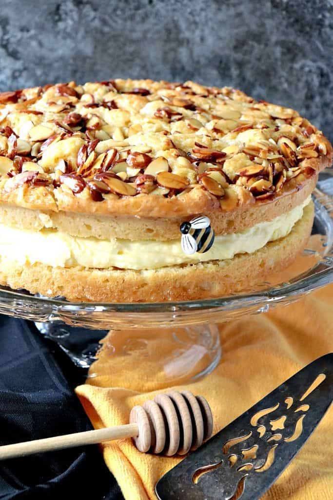 A side view closeup photo of a German bee sting cake on a glass cake plate with a gray background and a little faux bee.