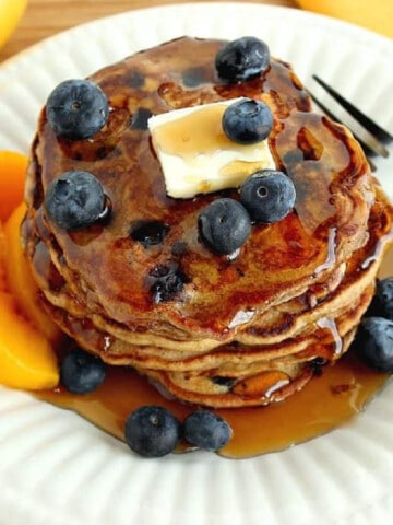 A stack of Blueberry Peach Pancakes on a white plate with a pat of butter and peaches on the side.