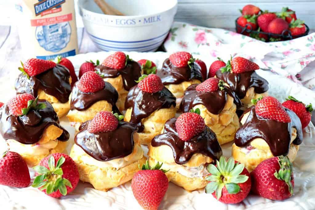 A plate of chocolate covered strawberry filled eclairs with a bowl and fresh strawberries as garnish.