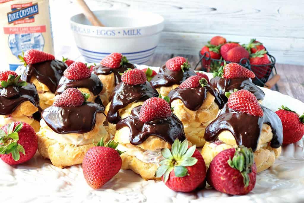 A horizontal photo of a tray filled with chocolate ganache covered strawberry filled cream puffs with fresh strawberries as garnish.