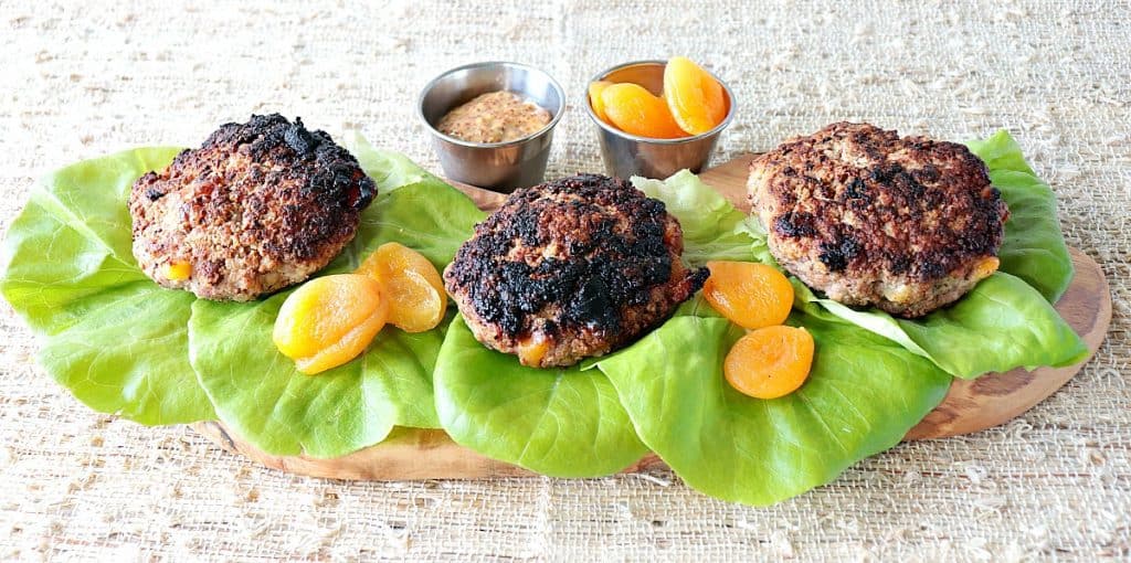 Three Pork Burgers with Dried Apricots (without buns) on a wooden board with lettuce leaves and dried apricots as garnish.