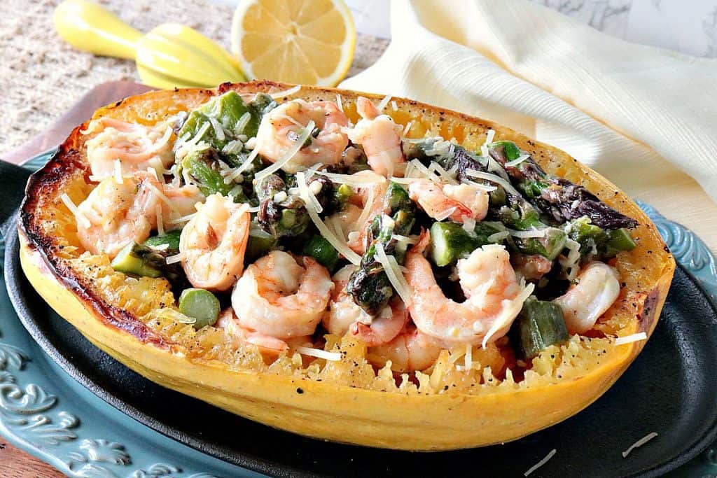 A roasted spaghetti squash on a plate with asparagus and shrimp with a lemon in the background.