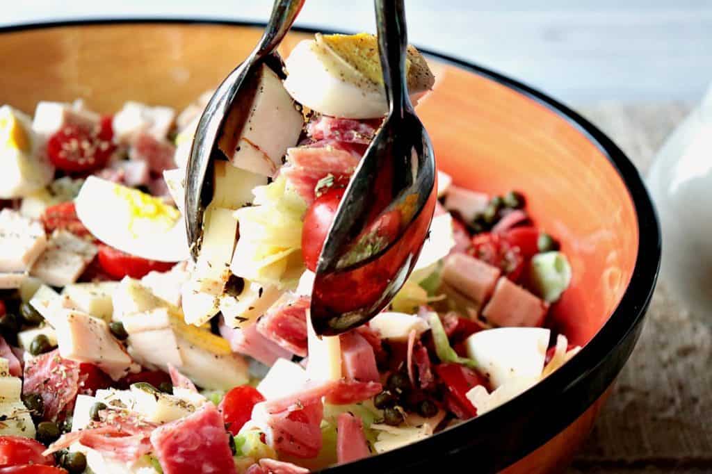 A pair of tongs serving up a chef's salad with meat, cheese, and lettuce.