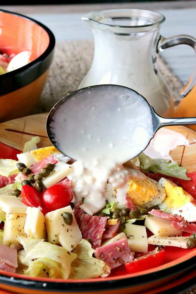 A vertical closeup photo of a chef's salad in a bowl with a serving spoon pouring on some blue cheese dressing.