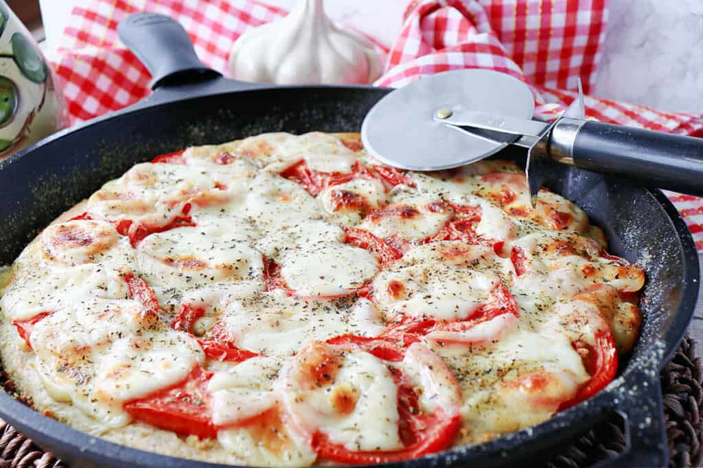 A closeup horizontal photo of a Deep Dish Garlic Lover's Pizza in a cast iron skillet with melted cheese and a pizza wheel.