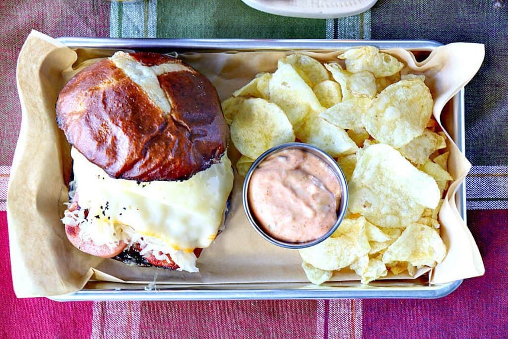 Overhead photo of a knockwurst sausage recipe on a pretzel roll with melted cheese, sauerkraut, chips, and sauce.