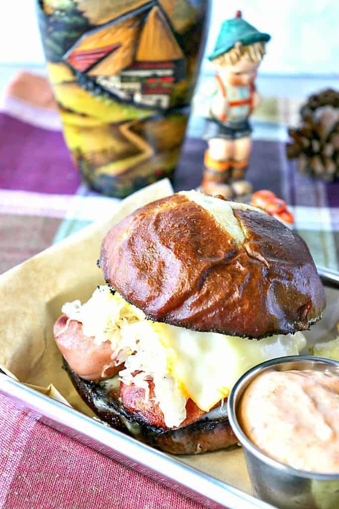 Vertical closeup photo of a knockwurst sandwich on a pretzel roll with melted cheese and a German vase in the background.