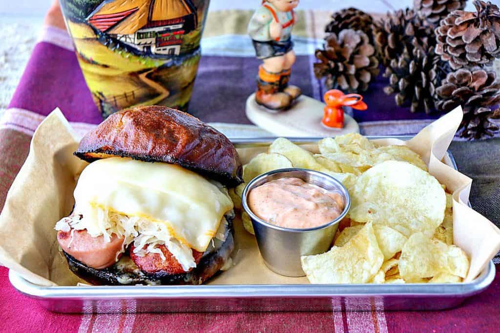 An open face German knockwurst sandwich with melted cheese and sauerkraut on a tray with chips.