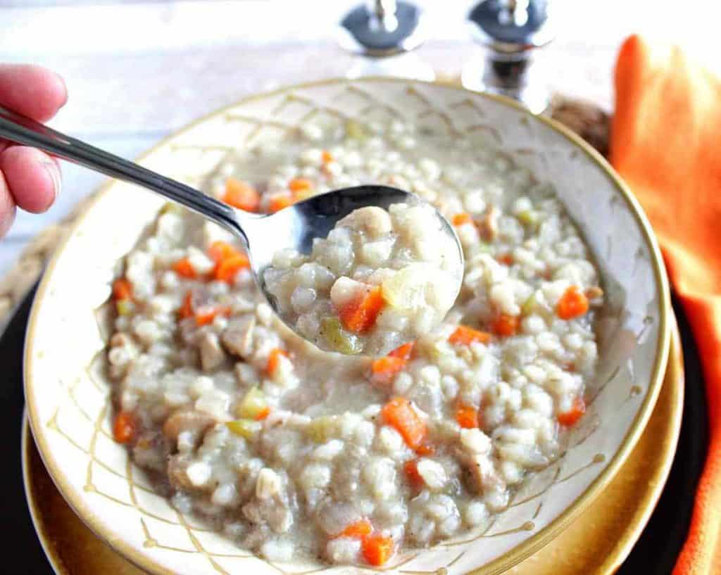 A closeup image of chicken and barley soup on a soup spoon with carrots, celery. Comfort food soup, stew, and chowders recipe roundup.