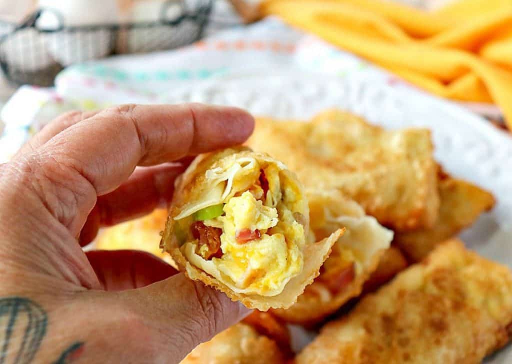 A closeup of a hand holding half of a breakfast egg roll filled with scrambled eggs, cheese, and bacon.