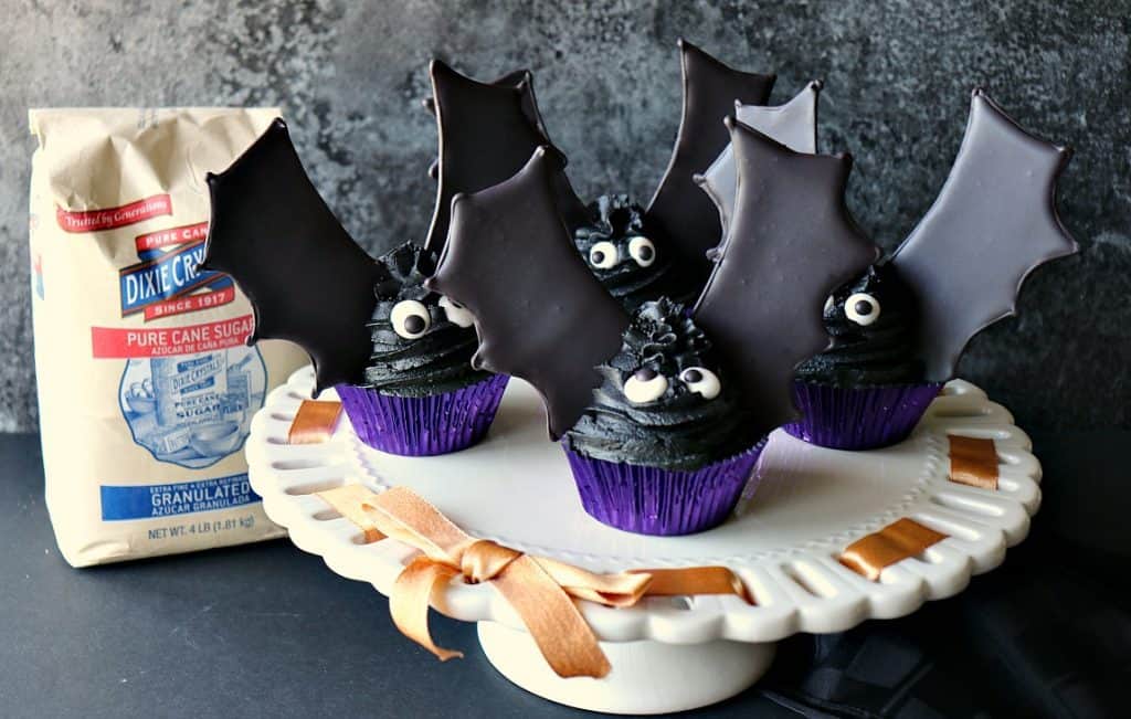 A flock of bat wing cupcakes on a white cake stand with orange ribbon