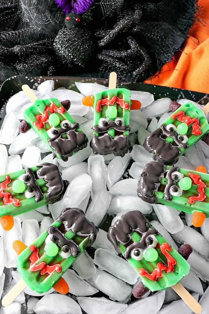 A bunch of frozen Frankenstein pudding pops on a tray with ice and orange and black napkins in the background.