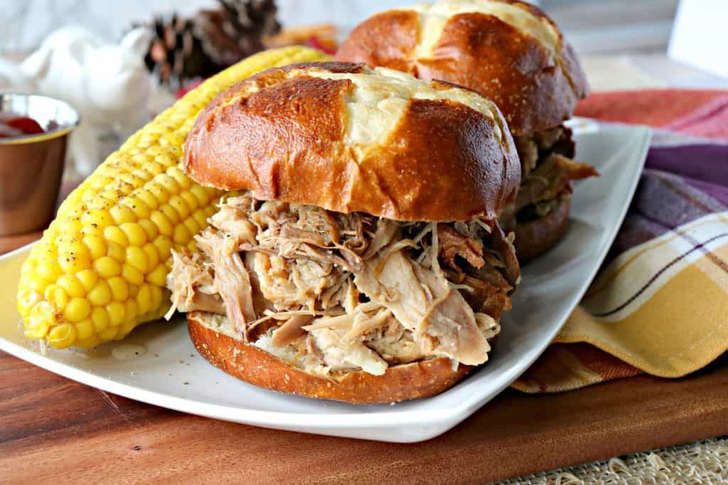 Two slow cooker pulled pork sandwiches on a plate with pretzel rolls and a corn on the cob.