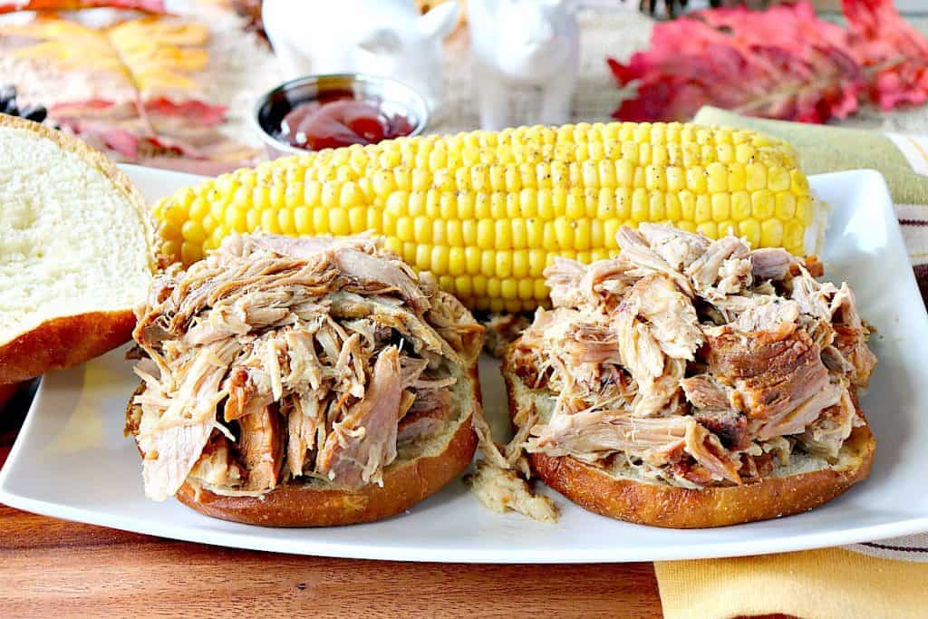 Two open faced slow cooker pulled pork sandwiches on a plate with a corn on the cob in the background.