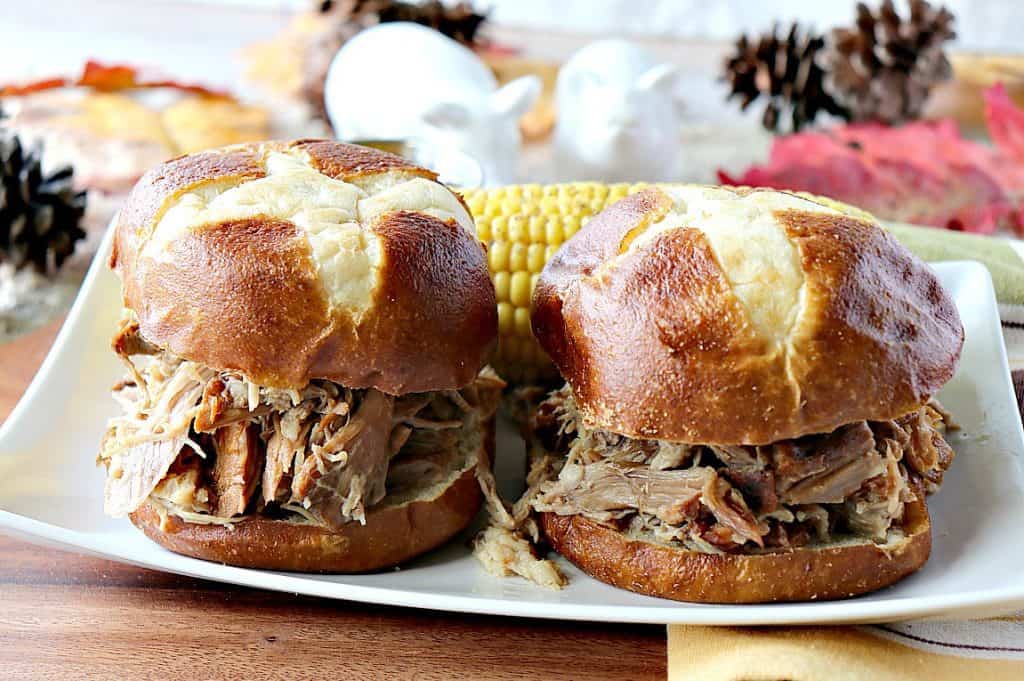 Two slow cooker pulled pork sandwiches on a white rectangle plate with acorns and a corn on the cob in the background