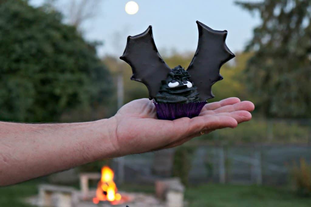 A hand holding up a bat wing cupcake outside with a full moon in the background and a fire in a firepit.