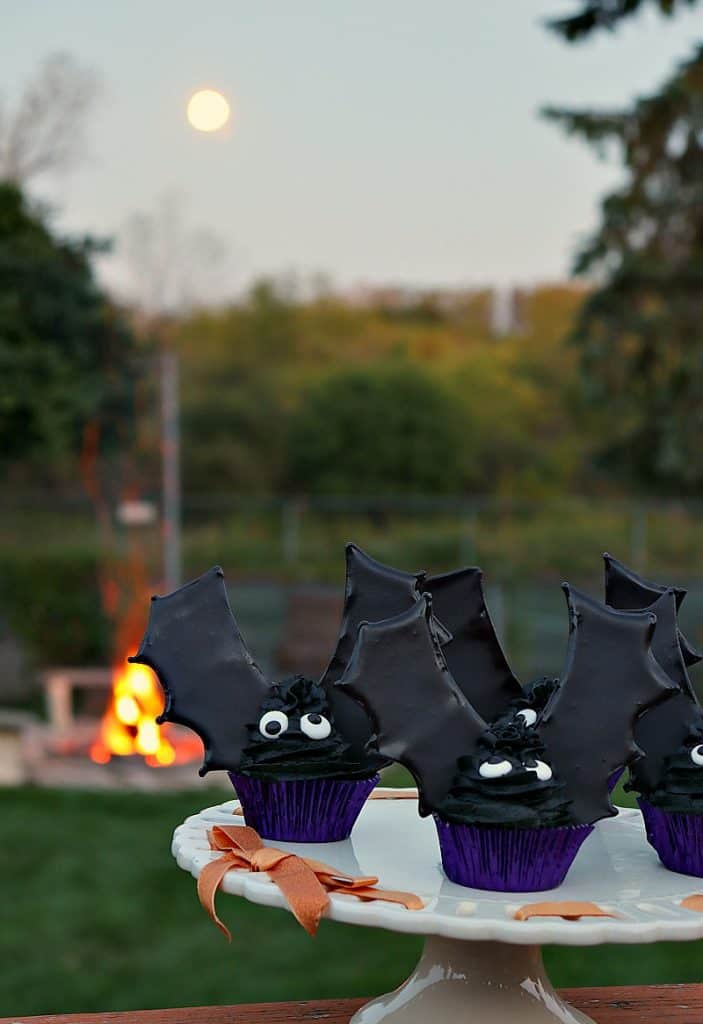 Vertical photo of bat wing cupcakes outside at dusk with a fire in the background and a full moon in the sky.