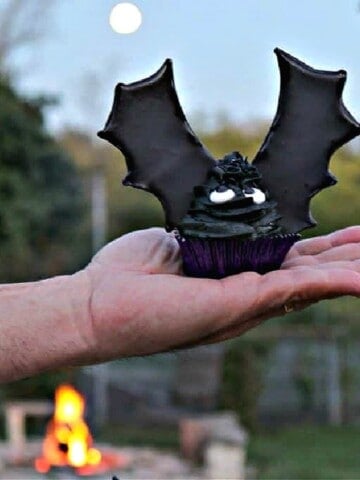 A hand holding a Batwing Cupcake with a moon and fire in the background.