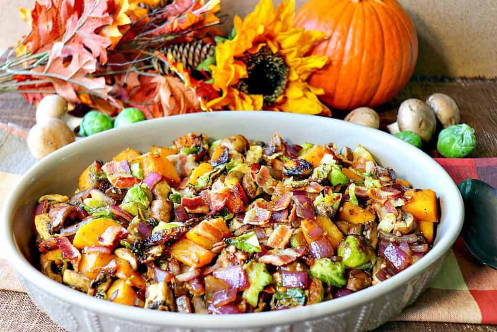 An oval casserole dish filled with colorful sauteed autumn vegetables of Brussels sprouts, mushrooms, butternut squash, and bacon. Pumpkin, mushrooms and a sunflower are in the background.