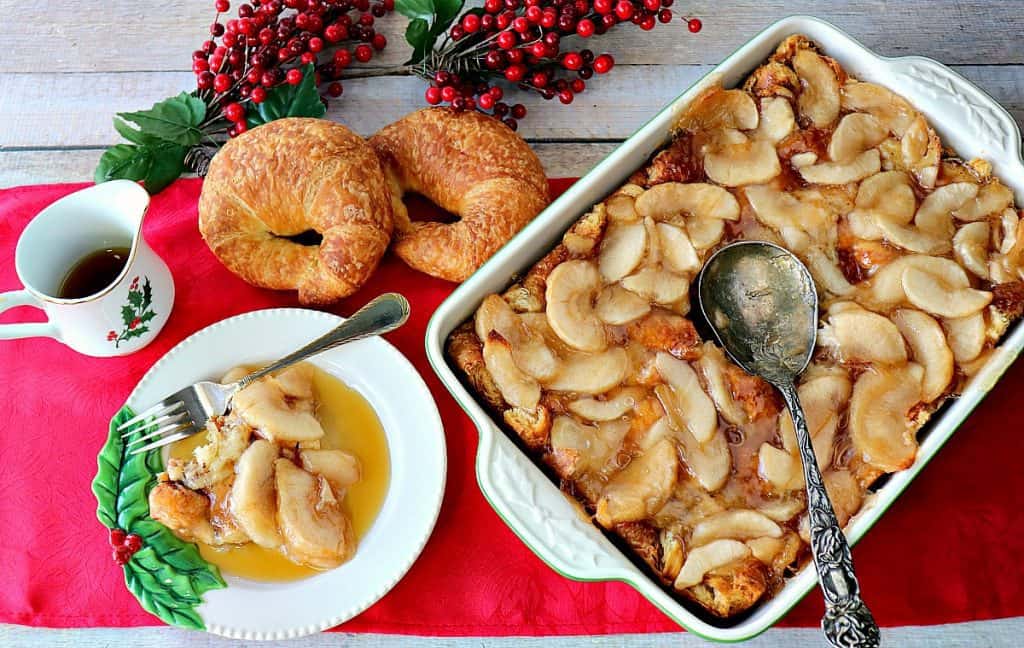 A horizontal overhead photo of a casserole dish filled with Apple Croissant Breakfast Bake along with two croissant rolls and a large serving spoon.