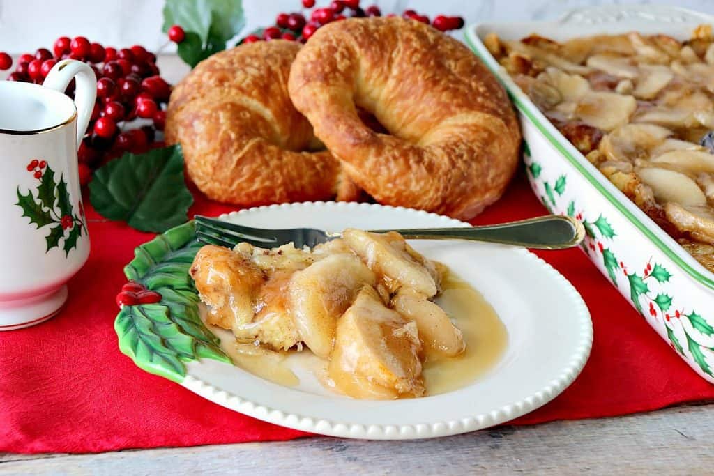 Horizontal photo of a serving of apple croissant breakfast bake on a pretty holiday plate with a red tablecloth and croissants in the background.