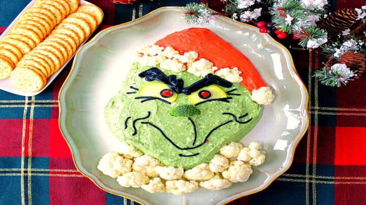 A fun Grinch Guacamole on a platter with crackers and some Christmas ornaments in the background.