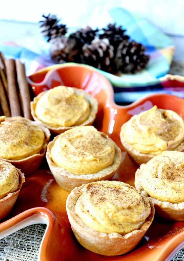 Vertical image of pumpkin cheesecake tartlets on an orange leaf plate for Thanksgiving dessert recipe roundup.