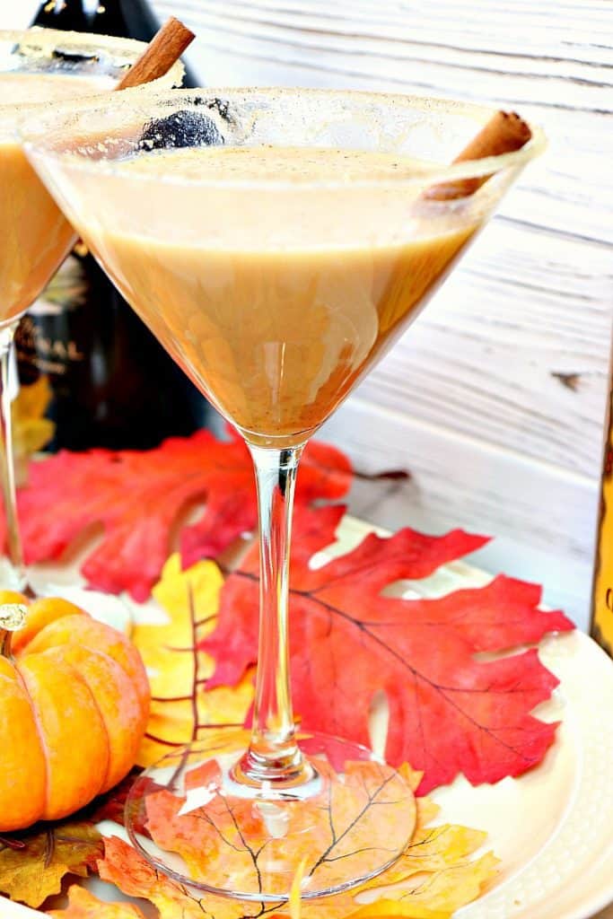 Vertical closeup of a pumpkin spice martini with autumn leaves and a cinnamon stick. New year's eve appetizer and drinks recipe roundup.