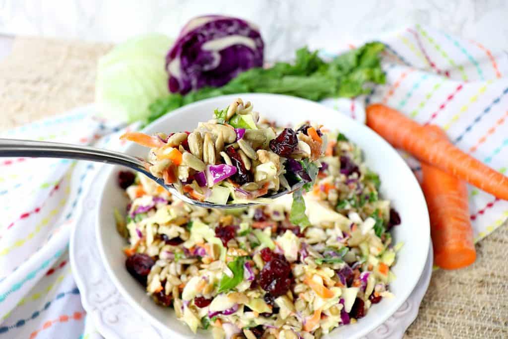 Colorful sunflower crunch kale and cabbage salad in a white bowl with carrots, red cabbage, and celery in the background. 