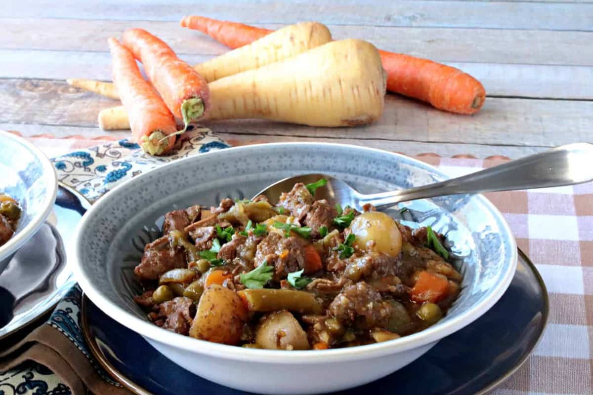 A horizontal photo of a hearty bowl of beef stew with carrots and parsnips in the background and a spoon in the bowl.