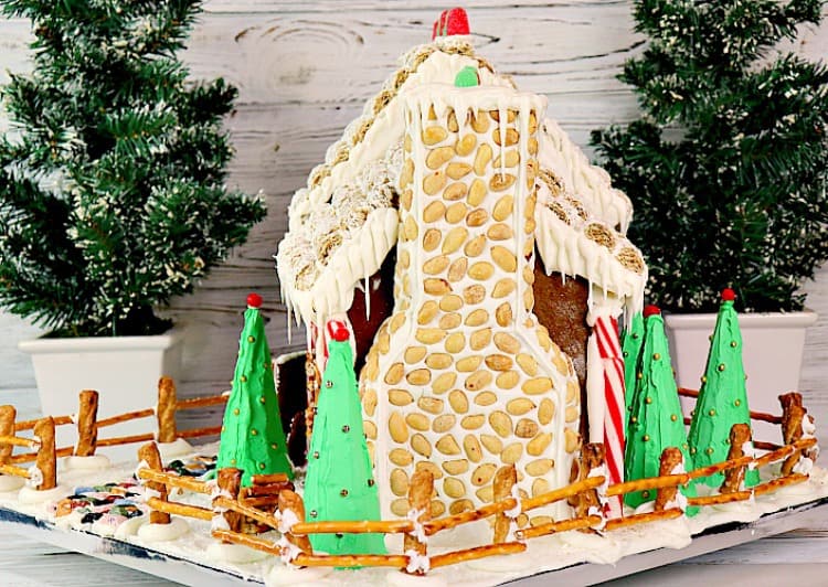 Side view of decorated gingerbread house with peanut and royal icing chimney, pretzel fence and sugar cone iced pine trees. - kudoskitchenbyrenee.com
