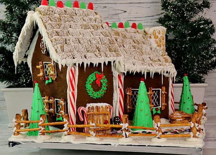 Horizontal photo of a gingerbread house with frosted wheat roof, candy cane sides, and a pretzel fence.