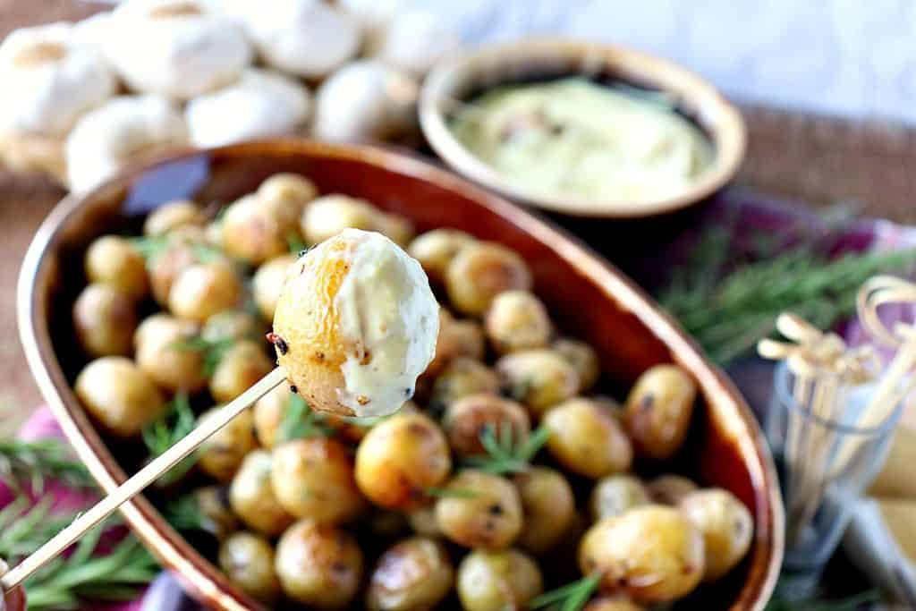 A closeup photo of one garlic herb roasted baby potato on a wooden pick and an entire bowl of potatoes in the background.