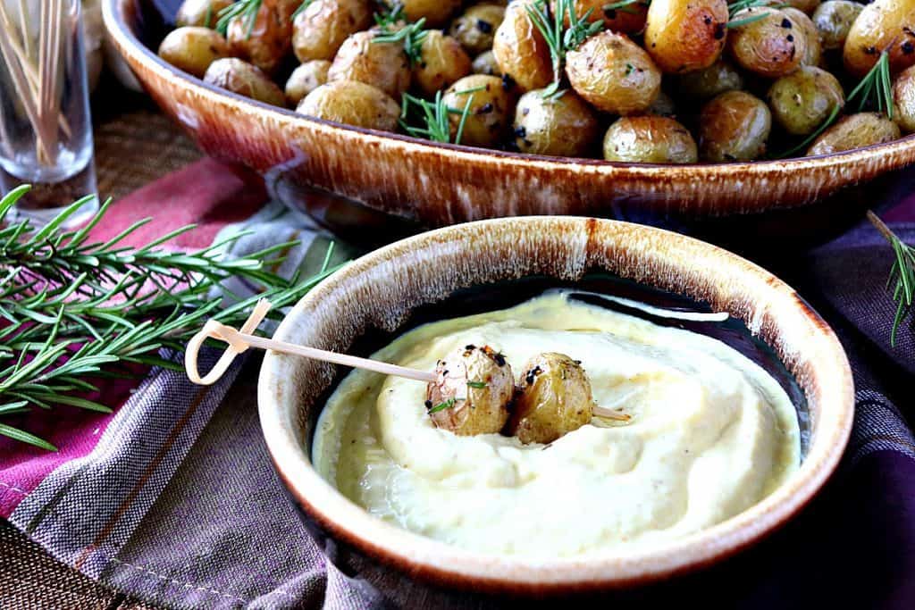 A small brown bowl of sauce with two baby garlic herb roasted potatoes on a wooden pick.