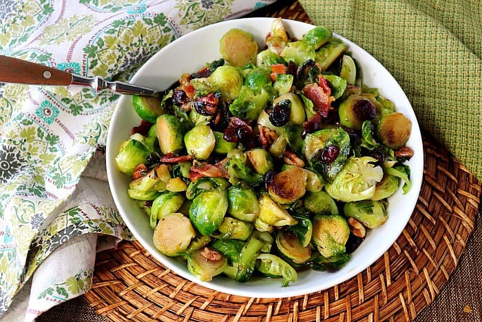 Sauteed Brussels sprouts in a white bowl on a rattan place mat and two napkins. Christmas dinner recipe roundup.