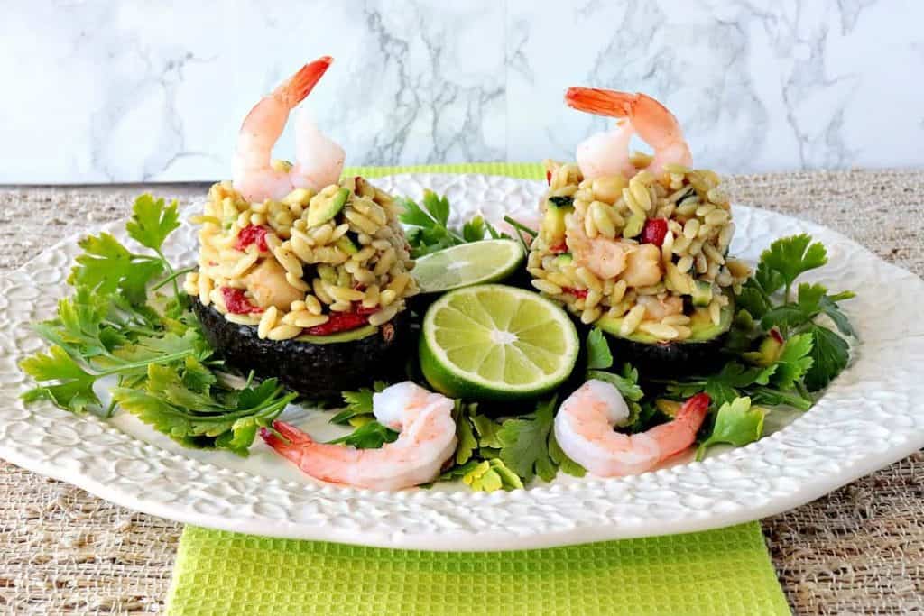 Two orzo stuffed avocados on a plate with lettuce, limes and shrimp. 