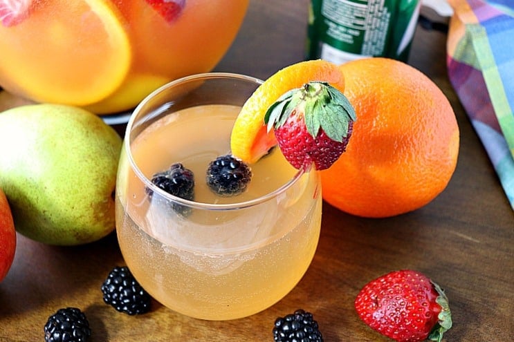 A horizontal photo of a colorful blackberry sangria spritzer in a wine glass with strawberries, blackberries, and oranges in the background.