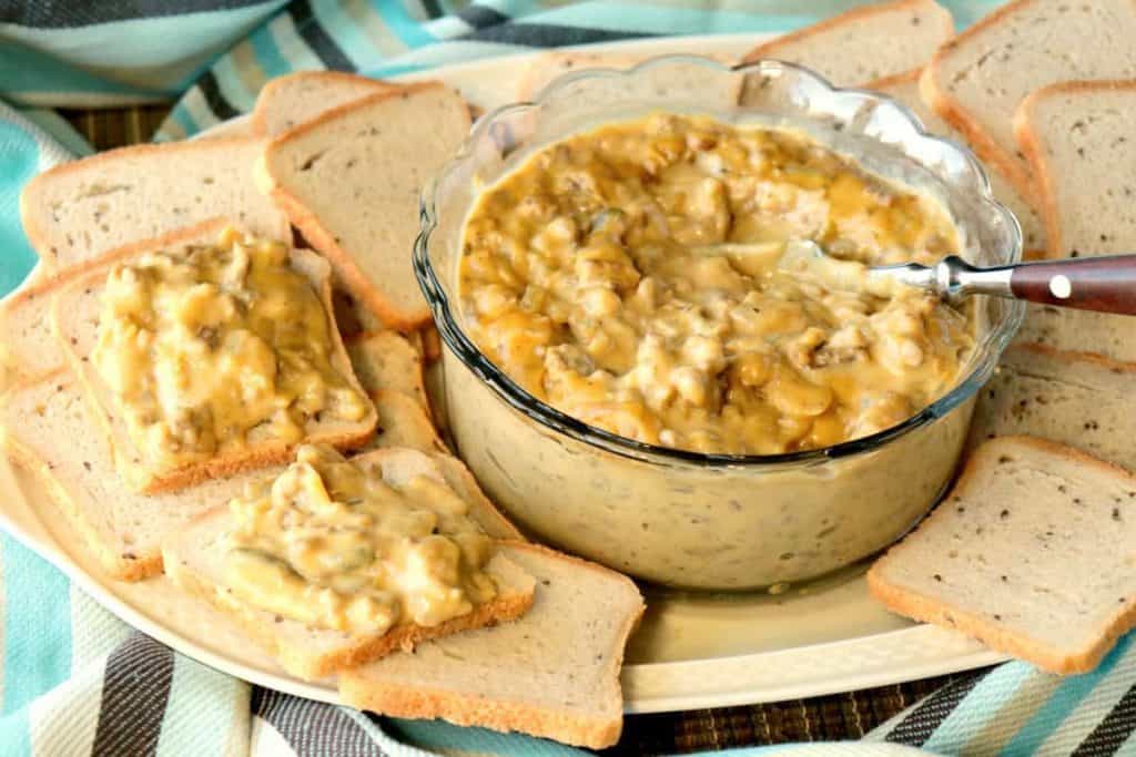 A glass bowl filled with patty melt dip along with some cocktail bread and a knife spreader.