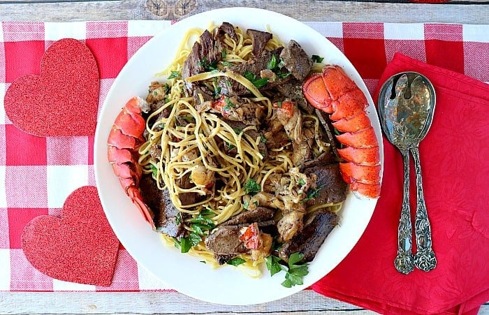 An overhead photo of a large pasta bowl filled with steak and lobster linguine with lobster tails on the side, a red and white checked table cloth, and serving utensils.