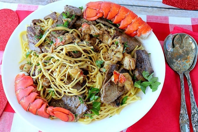 Steak and Lobster Linguine with two lobster tails on a plate with a red and white checkered tablecloth and a red napkin.