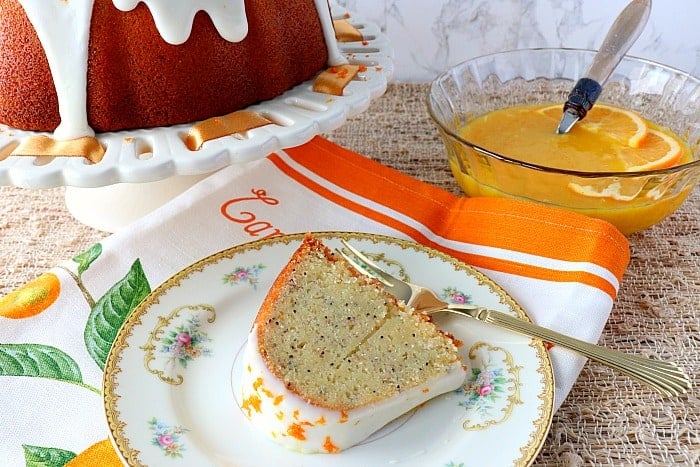 A slice of orange banana poppy seed bundt cake on a plate with a bowl of orange curd in the background.