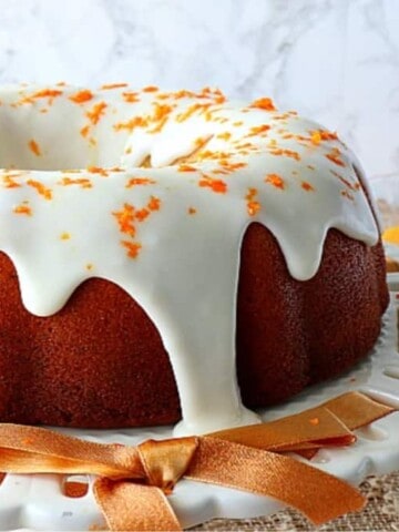 An Orange Bundt Cake on a cake stand with an orange ribbon and a bowl of orange curd in the background.