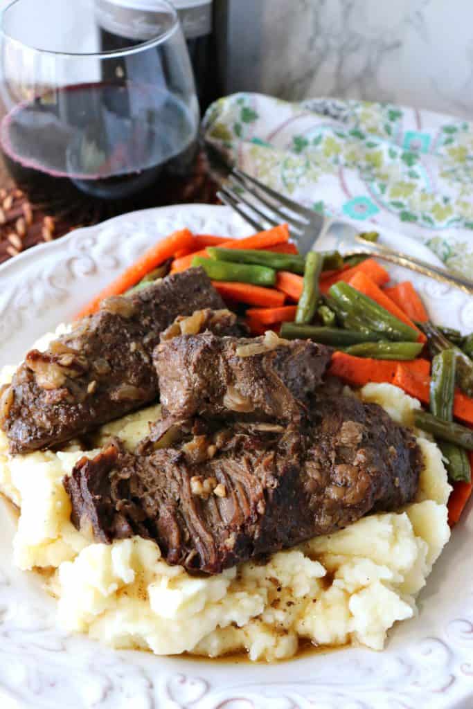 Beef short ribs over mashed potatoes on a plate with a glass of wine in the background.