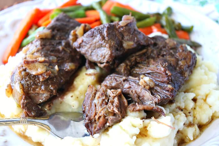 Closeup picture of short ribs over mashed potatoes on a white plate with carrots and green beans in the background.