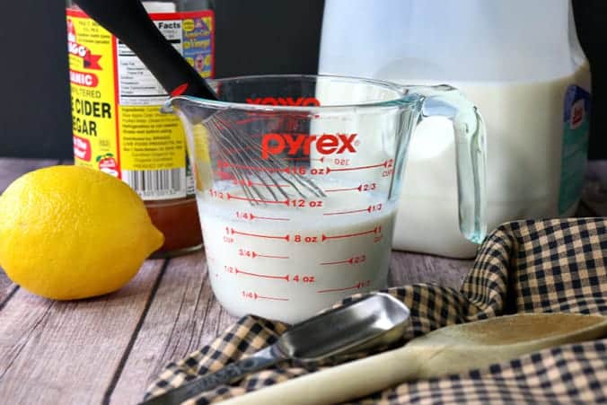 Buttermilk alternative inside a measuring cup with a lemon, apple cider, milk, spoons and a whisk surrounding it.