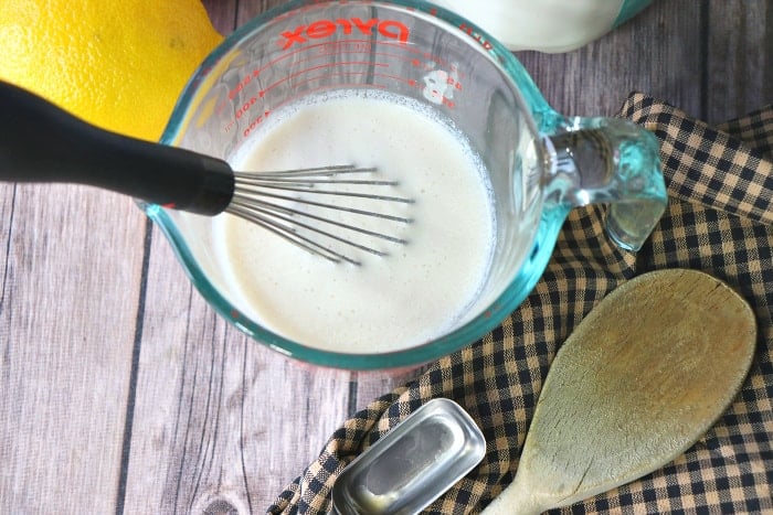 Closeup and overhead picture of homemade buttermilk in a measuring cup with a whisk, wooden spoon, and measuring spoon.