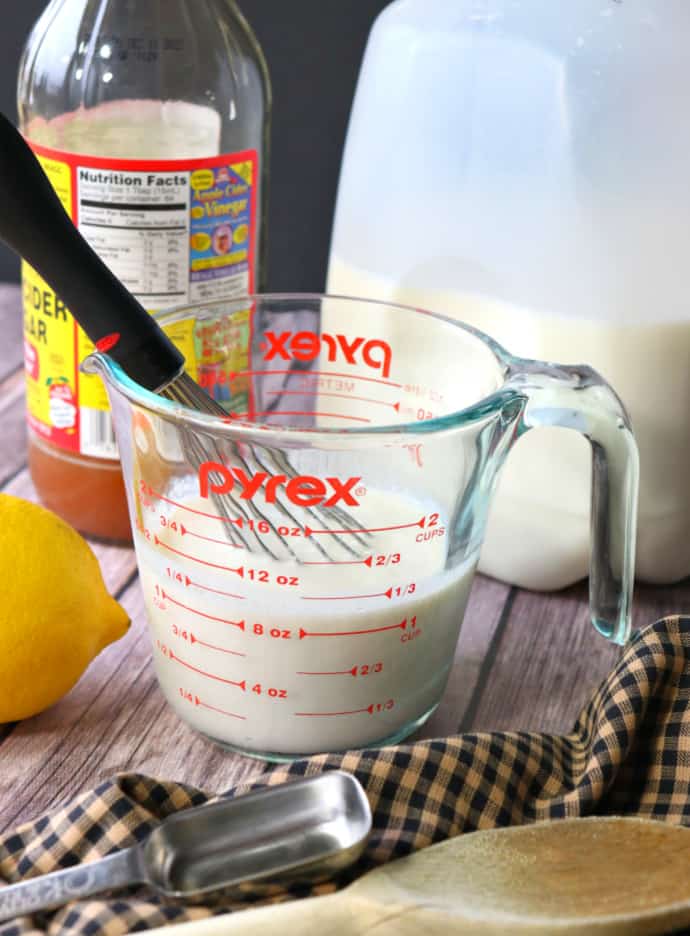 Vertical image of a measuring cup with buttermilk, whisk, vinegar, lemon, and milk in the background.