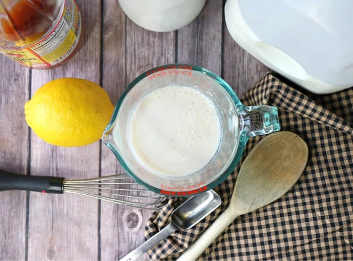 Straight overhead shot of buttermilk in a glass measuring cup with a tan and black check napkin, a wooden spoon, whisk, and a measuring spoon.