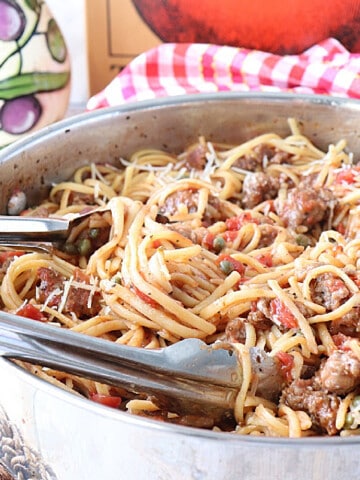 A silver skillet filled with Linguine with Sausage and Capers along with tongs.