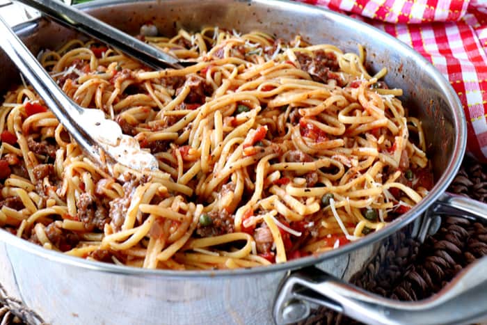 Pasta inside a large stainless steel pan with tongs and a red and white check napkin.